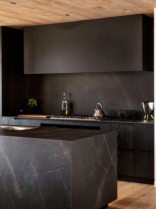 A dramatic, dark-toned contemporary kitchen featuring a large stone-clad island, matching stone splashback and benchtops, dark timber cabinetry, integrated appliances, and a warm timber-lined ceiling. Brass and metallic kitchen accessories provide subtle highlights.
