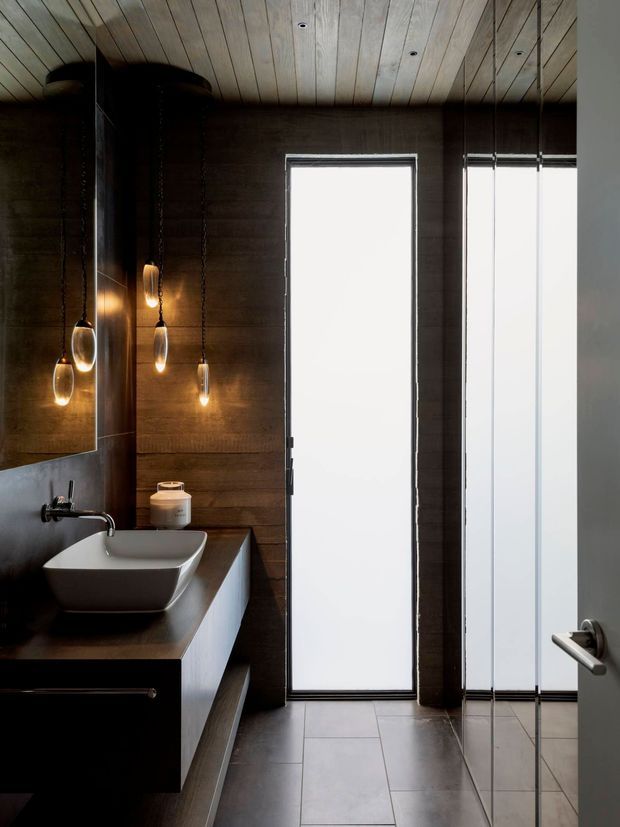 A dark, refined bathroom with a floating dark timber vanity, white vessel basin, wall-mounted tapware, large mirror, and slender pendant lights. The room features dark timber wall panelling, a light timber slatted ceiling, large frosted windows or doors, and large grey floor tiles.
