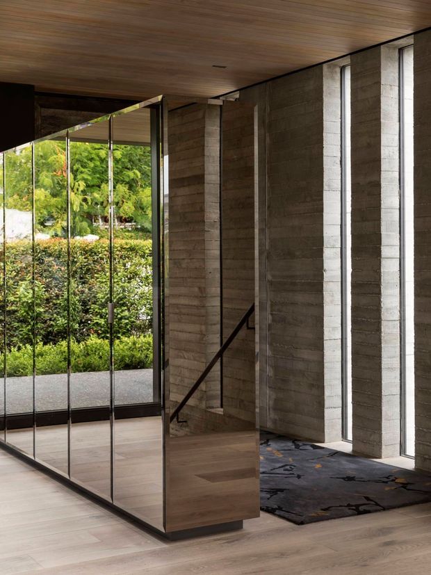 Interior view of a modern entry hall or circulation space with a dramatic cantilevered timber-clad stair enclosure, exposed horizontally textured concrete walls, slim dark metal framing, and full-height glazing overlooking lush garden planting. The space has warm timber flooring and ceiling, a dark abstract area rug, and a restrained material palette with strong natural light.
