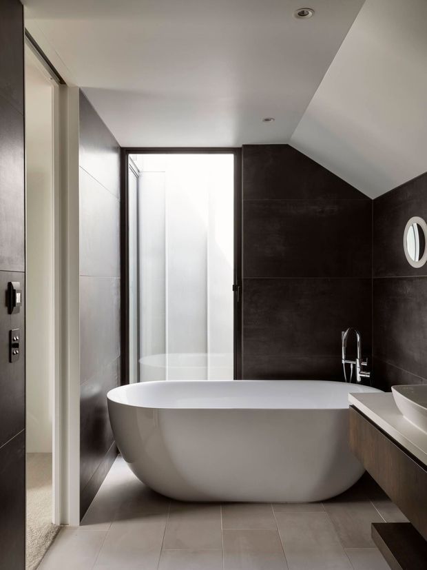 Contemporary bathroom with a freestanding white oval bathtub, dark charcoal wall tiles, pale grey floor tiles, a timber-toned floating vanity with white benchtop, vessel basin and chrome tapware. A frosted-glass window/door provides diffused natural light, while the sloping ceiling and circular wall mirror add distinctive architectural details.