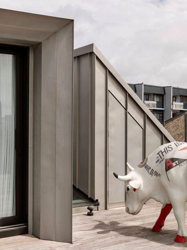 A contemporary rooftop terrace or balcony with a strong geometric architectural composition. The foreground shows a dark-framed glazed opening beside large projecting and angled grey metal-clad wall forms, with timber-toned decking underfoot. A sculptural painted white bull occupies the terrace, while multi-storey residential buildings are visible in the background beneath an overcast sky.