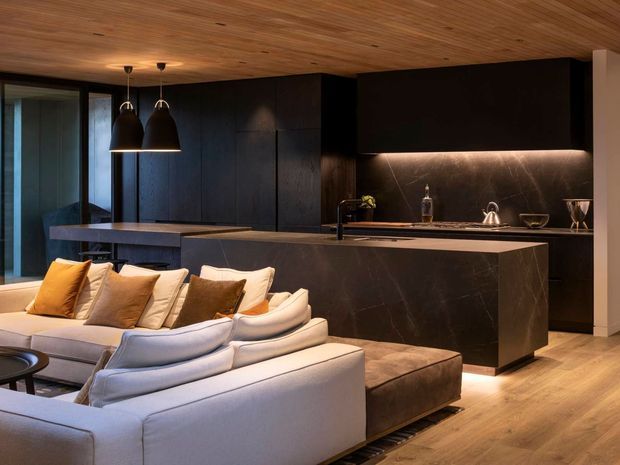 A sophisticated open-plan kitchen and living area with dark charcoal cabinetry, a large stone island and matching backsplash, warm timber ceiling and flooring, pendant lighting, and a light sectional sofa with neutral cushions in the foreground. The space has a refined, moody atmosphere with concealed under-cabinet lighting and dark integrated joinery.