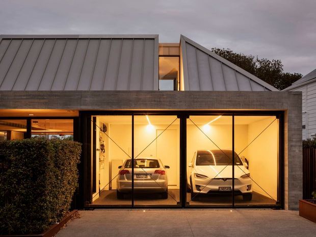 Twilight exterior view of a contemporary residential garage, with two cars visible behind full-height glazed garage doors. The facade combines dark horizontal cladding, folded standing-seam metal roof forms, black-framed glazing, warm interior lighting, and landscaped hedging.