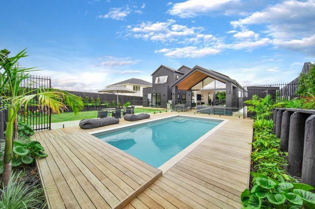A contemporary residential outdoor entertaining area with a rectangular swimming pool surrounded by a broad timber deck. The pool courtyard includes lounge seating, tropical planting, dark fencing, and a covered pavilion with a pitched gable roof, glazed walls, and warm timber soffit detailing. A modern grey house is visible beyond.
