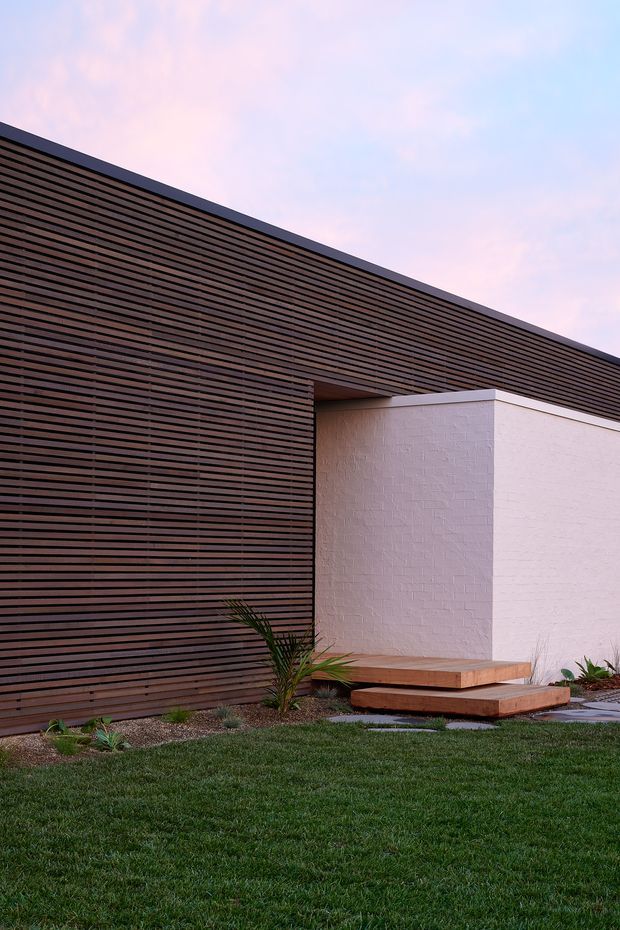 A minimalist modern building facade with a dramatic low-pitched or flat roofline, clad in closely spaced dark timber slats. A projecting white textured masonry volume creates a strong geometric contrast, with timber steps and a simple landscaped garden at its base. The image is photographed externally at dusk beneath a softly coloured sky.
