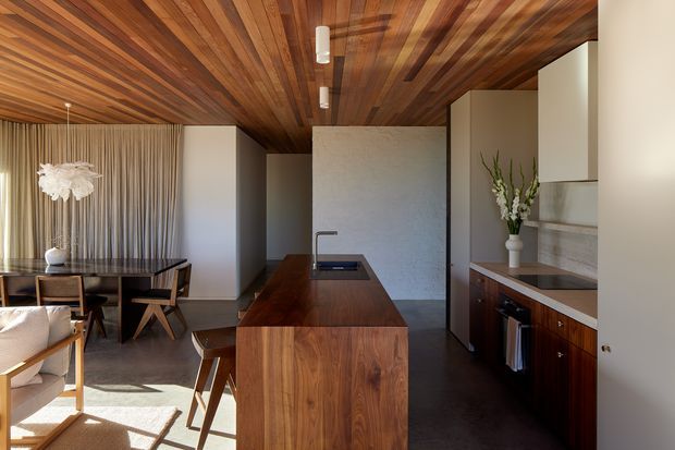 Contemporary open-plan kitchen and dining area with a substantial dark timber island, integrated cooktop, dark cabinetry, and pale stone or plaster finishes. The room features a dramatic warm timber-lined ceiling, full-height curtains, sculptural pendant lighting, timber dining furniture, and strong natural light.