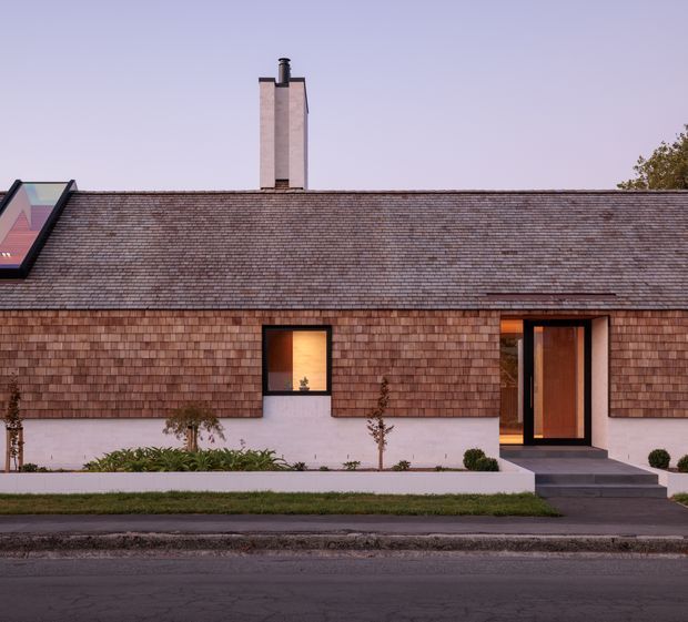 A low, single-storey residential facade viewed from the street, with a long pitched roof clad in small brown timber shingles, matching shingle-clad walls, a pale rendered base, and a prominent brick chimney. A dark-framed roof window is visible at the left, while the central window glows warmly and the recessed entry has a tall timber door with dark framing and shallow steps. Minimal landscaping and a paved street occupy the foreground.
