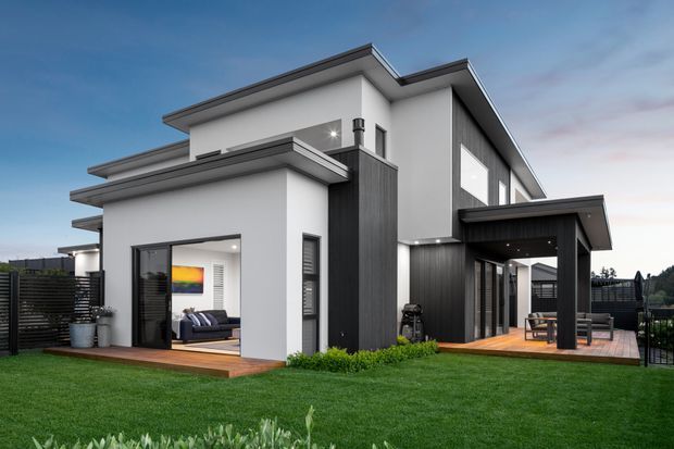 Exterior photograph of a contemporary multi-level home with crisp white rendered walls, dark vertical cladding, flat and low-pitched roof planes, large glazed openings, and timber decking. The landscaped lawn and covered outdoor deck are prominent, with glimpses of an interior living area through the open sliding doors.