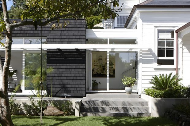A bright residential exterior showing a contemporary glazed extension attached to a white traditional timber-clad house. The extension features full-height glass doors and windows, a dark shingle-clad wall, white parapet-like roof detailing, and broad concrete steps leading into a landscaped front garden. The original house has a pitched roof, white weatherboard cladding, and divided-light windows.