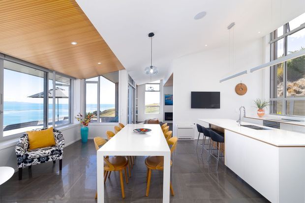 Bright open-plan kitchen and dining area with a long white island, integrated sink, timber stools, and a separate dining table. Floor-to-ceiling glazing overlooks a blue coastal body of water, while a timber-lined ceiling zone, polished dark flooring, white cabinetry, pendant lights, and a compact living area create a clean contemporary interior.