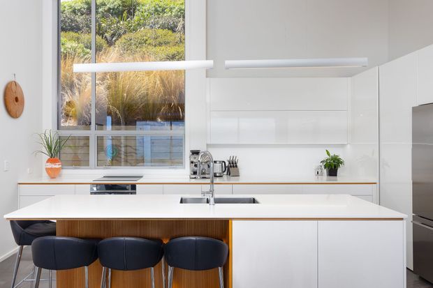 Bright contemporary kitchen with a large white island, integrated sink and tap, white full-height cabinetry, timber island detailing, dark upholstered bar stools, stainless-steel appliances, and linear pendant lighting. Floor-to-ceiling glazing provides views to landscaped planting outside.