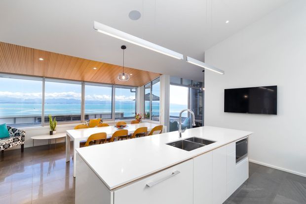 Bright contemporary open-plan kitchen and dining area with a large white island, integrated sink and appliances, timber dining table and chairs, expansive floor-to-ceiling glazing, and a warm timber-lined sloped ceiling. A waterfront landscape is visible beyond the windows.