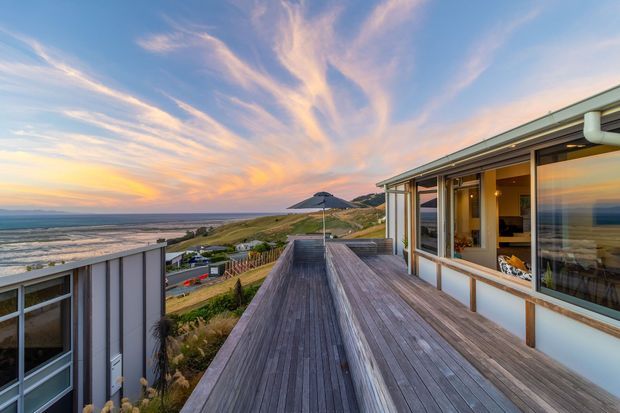 A coastal residential deck at sunset, with expansive weathered timber decking, a low contemporary home with large glazed openings and pale vertical cladding, and a seaside landscape with waves and dunes beyond. The image emphasizes the outdoor terrace, ocean outlook, and warm evening sky.