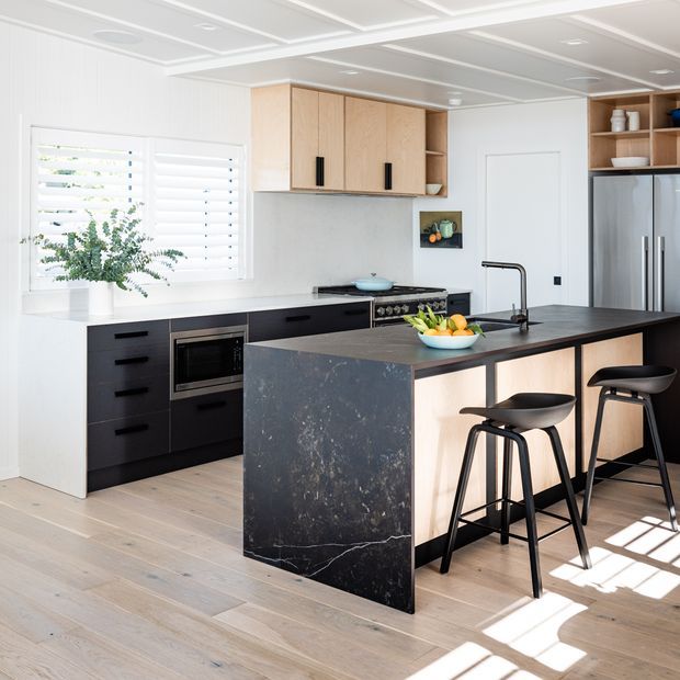 Bright contemporary kitchen with a large island featuring a dark stone waterfall benchtop, light timber cabinetry, black lower cabinetry, integrated appliances, and black bar stools. White walls, plantation shutters, pale timber flooring, and a coffered ceiling create a clean, airy interior.