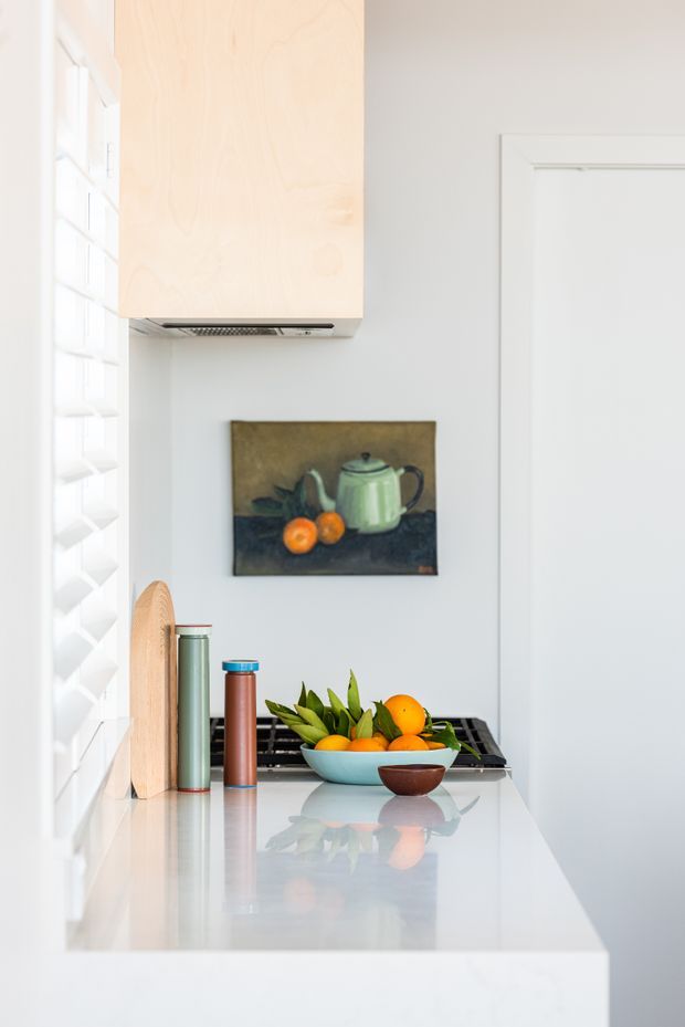 Bright, compact kitchen vignette with a glossy white benchtop, pale timber overhead cabinetry, white walls and trim, and a louvred window on the left. A fruit bowl, thermoses, cutting board and artwork sit on the counter, creating a clean, lightly styled residential interior.