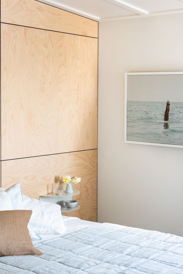 A bright, calm bedroom corner featuring a bed with pale blue and white bedding, a warm plywood feature wall with integrated panel lines, a small round bedside table, and neutral painted walls. A framed coastal photograph hangs above the bed area, while recessed ceiling detailing adds a refined contemporary finish.