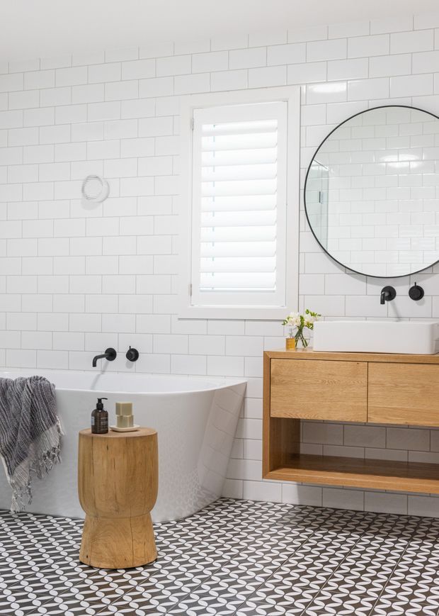 Bright contemporary bathroom featuring white horizontal subway-tile walls, a freestanding white bathtub, wall-mounted timber vanity with open shelf, round black-framed mirror, matte black tapware, a shuttered window, and patterned black-and-white floor tiles. Warm timber accents and monochrome finishes create a clean, refined look.
