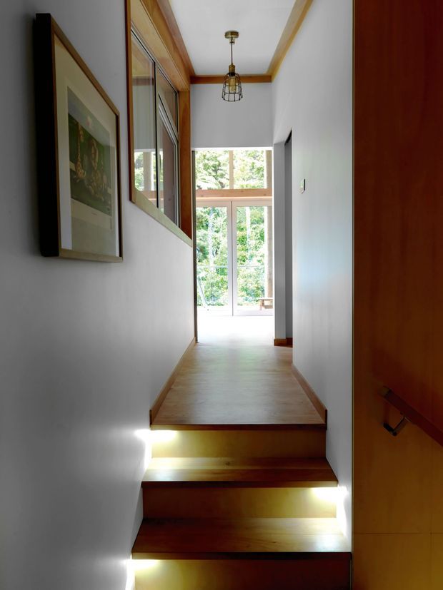 A narrow interior hallway with a short flight of timber stairs leading to a bright landing and full-height glazed opening. White walls are paired with warm timber flooring, stair treads, door surfaces, skirting, and ceiling trim. Framed artwork and interior glazing line the left wall, while recessed lighting illuminates the stair risers and a pendant light hangs above.