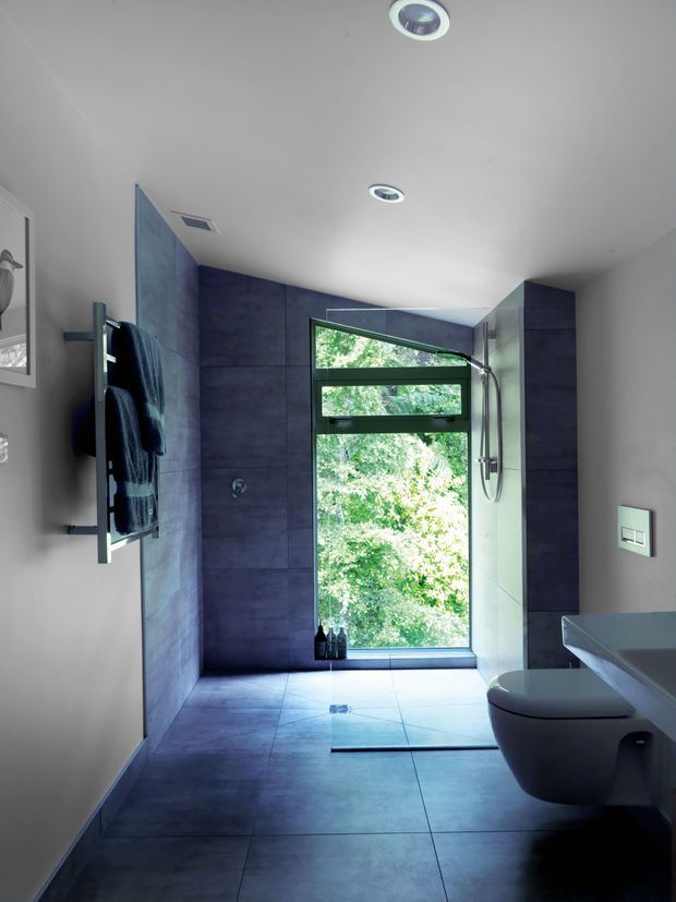 Contemporary bathroom with large-format dark blue-grey tiled walls and floor, a walk-in shower with rainfall and handheld fittings, wall-hung toilet, floating vanity, heated towel rail, recessed ceiling lights, and a tall window overlooking greenery. The bathroom has a clean, minimalist feel with strong natural light and dark-framed glazing.
