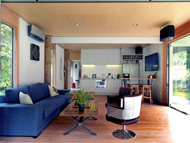 Open-plan living room and kitchen with a blue sectional sofa, timber coffee table, lounge chair, compact kitchen cabinetry and island bench. The room has extensive glazing, warm timber flooring and ceiling lining, integrated ceiling lighting, and a small dining or bar area. A leafy outdoor setting is visible through the windows and sliding doors.