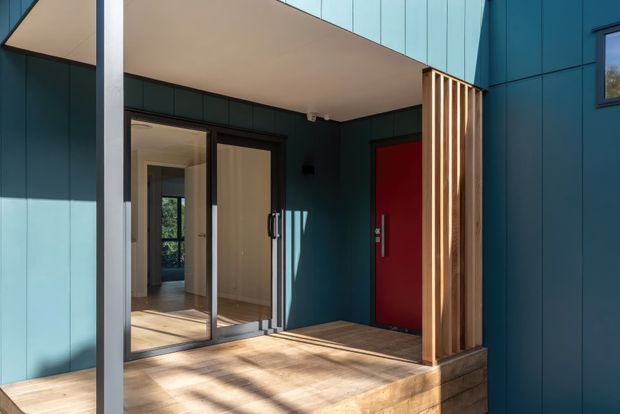 Contemporary residential entry facade with deep teal vertical cladding, a bold red front door, large glazed entry doors, pale timber decking, and a projecting flat-roof canopy. A vertical timber batten screen frames the entrance and adds warm contrast to the dark exterior. The view is from outside and focuses on the covered front porch and building envelope.