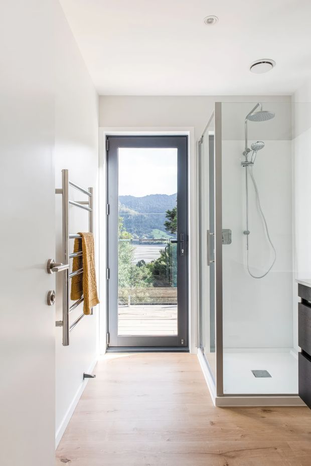 A bright, narrow contemporary bathroom with a walk-in glass shower on the right, rainfall and handheld shower fixtures, a wall-mounted heated towel rail with a mustard towel, pale timber-look flooring, white walls and ceiling, and a dark-framed glazed external door opening to a timber deck and leafy mountain view.