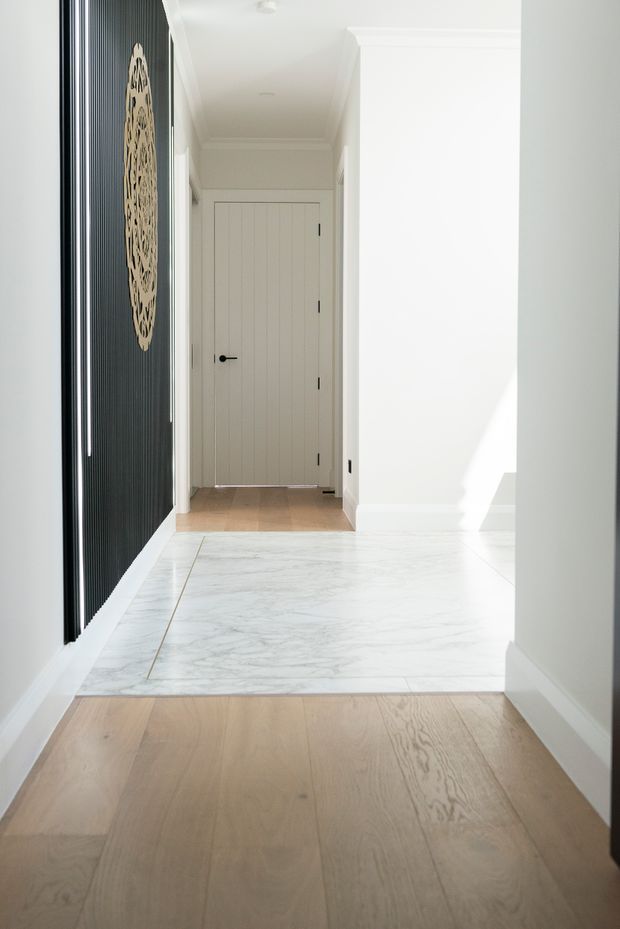 Bright residential hallway or entry passage with pale timber flooring in the foreground and a large white marble-look tiled section leading to a white vertically panelled door. The walls and ceiling are white with traditional cornice and skirting details. A dramatic black vertically slatted feature wall with a light decorative circular artwork provides contrast on the left. Natural sunlight enters from the right, creating a clean, refined interior.