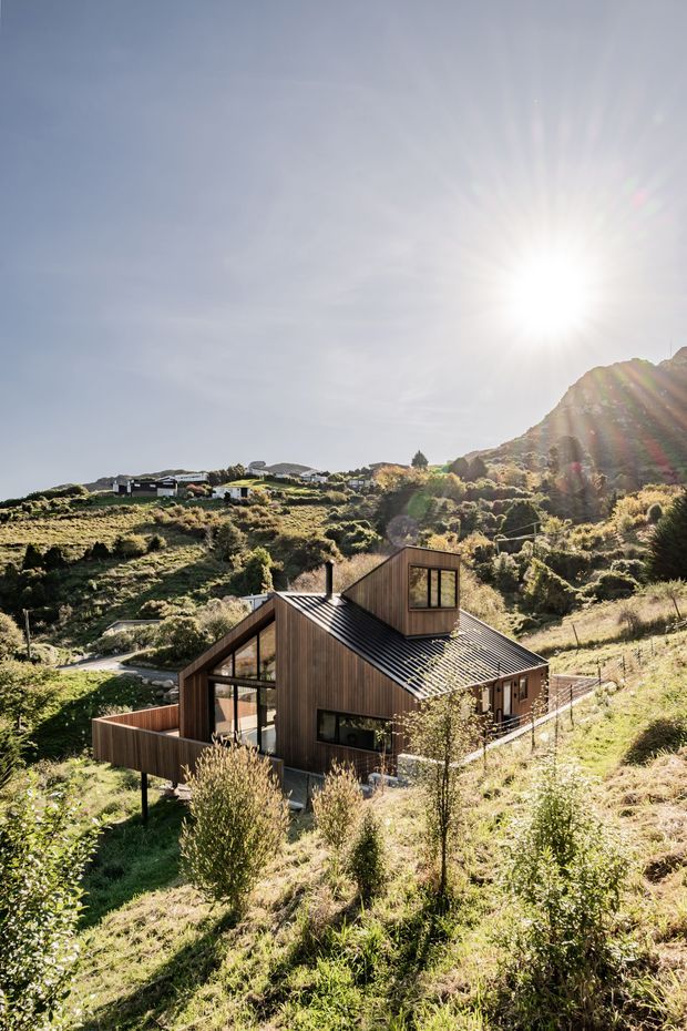 Exterior view of a compact contemporary timber-clad house set into a steep, sunlit rural or alpine hillside. The home has a strongly pitched dark metal roof, projecting upper dormer-like volume, extensive glazing, and timber deck/balcony areas. Surrounding scrub-covered slopes and a rugged rocky hill form the setting.