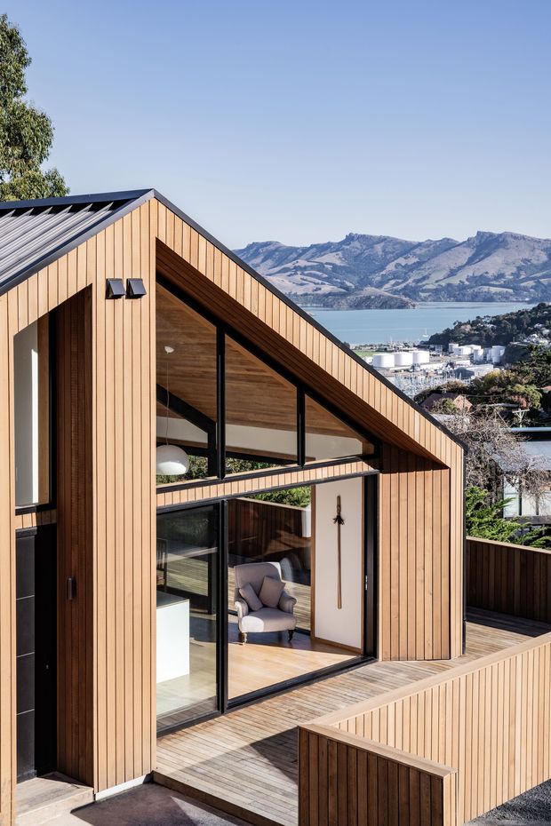 Exterior architectural photograph of a contemporary timber-clad house with a steep gabled roof, large black-framed glazed openings, and a timber deck and privacy balustrade. The elevated house overlooks a bay, harbour-side development, and mountainous terrain. Warm vertical timber cladding and exposed timber soffits contrast with dark metal framing and a clear blue sky.