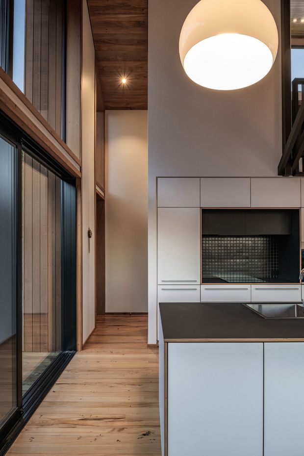 A contemporary kitchen viewed from a narrow circulation space, featuring pale flat-panel cabinetry, a dark benchtop, integrated sink, and a dark tiled backsplash niche. The room has warm timber flooring and a timber-lined ceiling, with tall black-framed glazing along the left side. A large spherical pendant light hangs above, and a high-level void or stair structure is visible at the right edge.