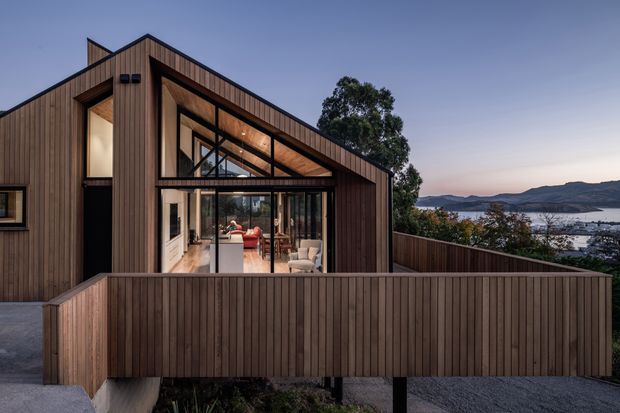 Exterior architectural photograph of a contemporary timber-clad home at dusk. The house has a steep asymmetrical gabled roof, exposed timber soffits and rafters, expansive black-framed glazing, and a raised timber deck enclosed by a vertical timber privacy screen. A warmly lit living area is visible through the glazing, with a coastal bay and hills in the background.
