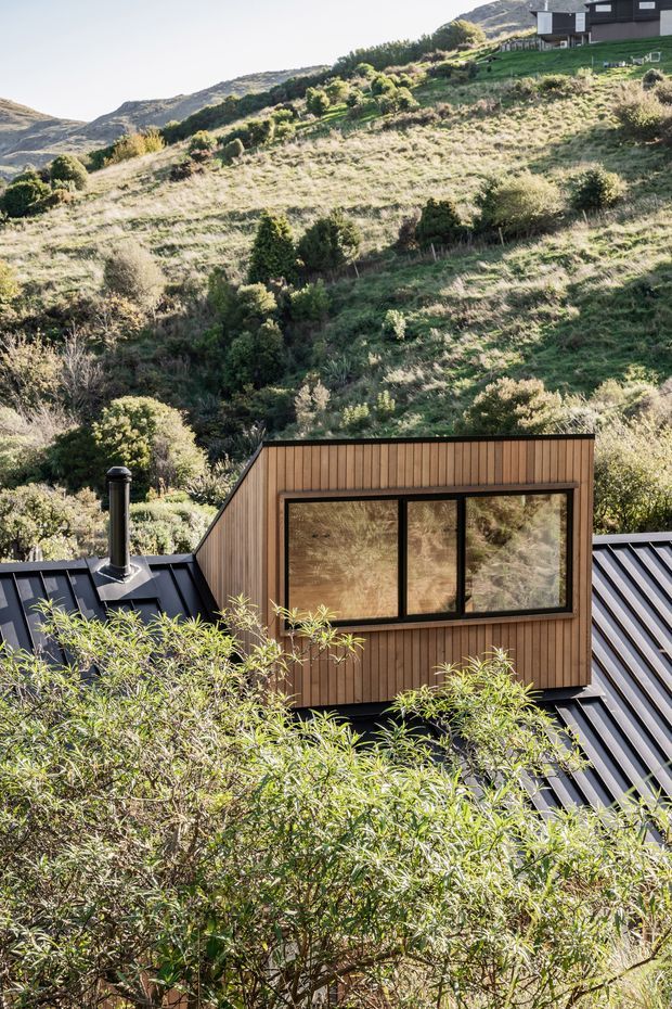 Exterior architectural photograph of a compact contemporary dwelling set into a steep, vegetated mountain hillside. The foreground shows a dark standing-seam metal roof with a projecting timber-clad dormer or upper volume, large dark-framed windows, and a black flue. A second dark-clad building is visible higher on the slope. The setting is rugged, elevated and alpine or mountainous, with dense native vegetation surrounding the home.