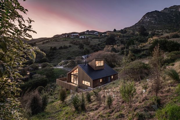 A compact dark-clad house nestled into a steep, rugged hillside at dusk. The dwelling has a pitched metal roof with a projecting roof dormer, large warm-lit timber-framed windows, and a small fenced terrace or courtyard. Dense native-looking vegetation and surrounding mountain terrain create a secluded rural or alpine setting.