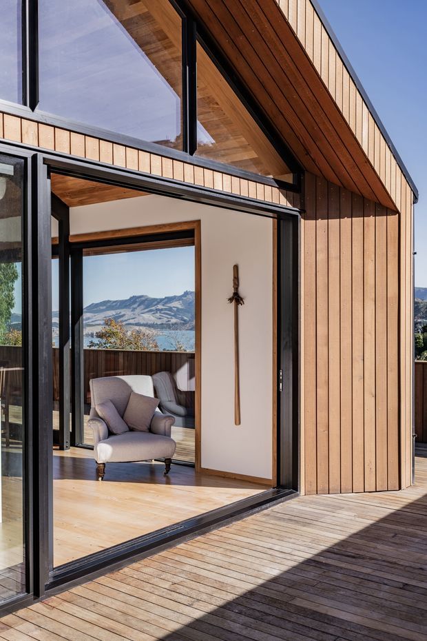 A close exterior view of a contemporary timber-clad residence opening onto a broad timber deck. Large black-framed glazed sliding or folding doors reveal a bright sitting area with an upholstered armchair and timber flooring. The building has strongly expressed warm timber soffits and vertical wall cladding, with a mountain and lake landscape visible through the interior glazing.