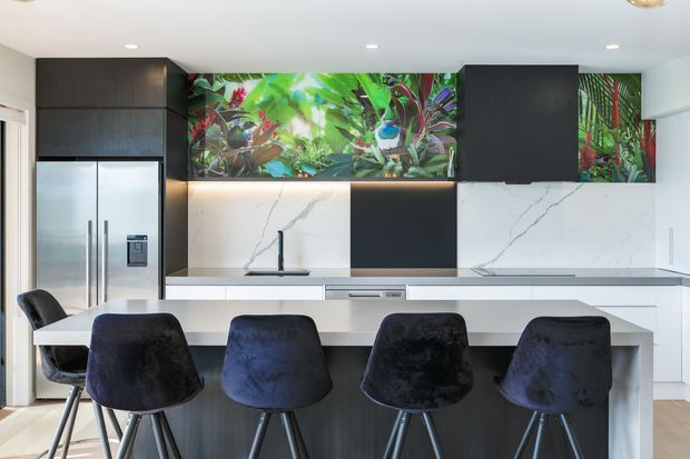A contemporary kitchen with a large white island and four dark upholstered bar stools. The space features white veined stone surfaces, matte dark cabinetry, integrated stainless-steel appliances, under-cabinet lighting, and a vivid tropical botanical splashback artwork or panel.