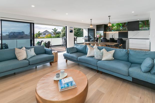 Bright open-plan living area with a large blue sectional sofa and matching armchair arranged around a round timber coffee table. The space has light timber flooring, white walls and ceiling, expansive sliding glass doors opening to a balcony or outdoor area, and a contemporary kitchen with dark cabinetry, white joinery, pendant lights, and a colourful botanical artwork backsplash.
