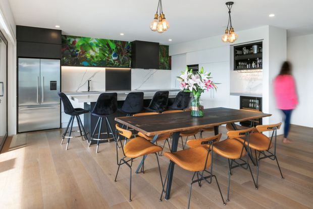 Bright open-plan kitchen and dining area with a large dark timber dining table, sculptural wooden chairs, a contemporary black kitchen island with stools, stainless-steel double-door refrigerator, dark cabinetry, white marble-look splashback, and a built-in display and bar area. Decorative pendant lights and a colourful botanical mural add visual interest.