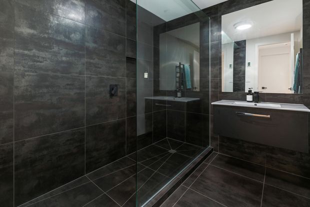 A contemporary bathroom with a large frameless glass walk-in shower, dark charcoal stone-look wall and floor tiles, and a floating dark vanity with a white benchtop and integrated basin. A large mirror, recessed shower fittings, and minimalist black fixtures create a sleek, moody interior.