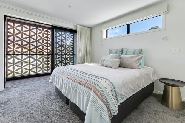 Bright contemporary bedroom with a low-profile upholstered bed dressed in layered white and pale blue-grey bedding. The room has soft grey carpet, white walls and ceiling, sheer curtains, and a long horizontal clerestory window. Full-height glazed doors feature a prominent geometric patterned privacy screen, while a small metallic side table sits beside the bed.