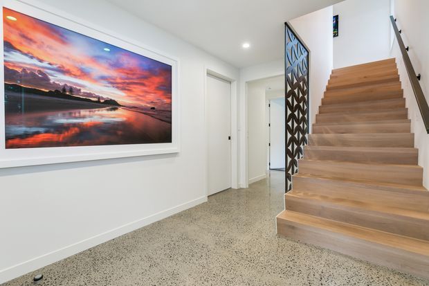 Bright contemporary residential hallway and stairway with white walls, a polished speckled terrazzo floor, and a broad light timber staircase. A decorative dark geometric screen forms the stair balustrade, while a large framed landscape artwork provides a colourful focal point.