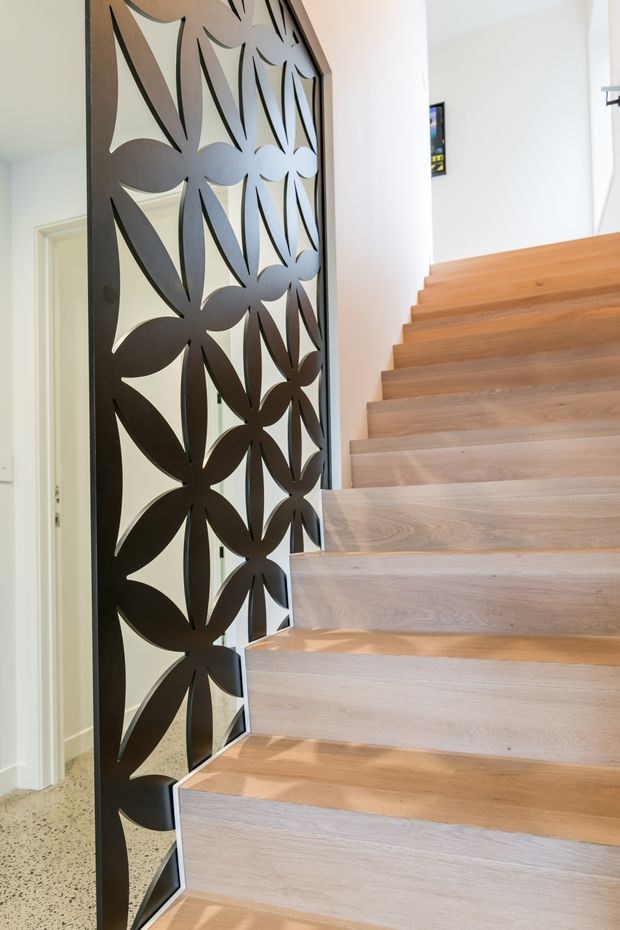 Bright contemporary stairway with light timber floating-style treads and risers, paired with a dark decorative metal screen featuring a repeating organic floral cut-out pattern. White walls and a terrazzo-like floor are visible beside the stair, with a doorway beneath or adjacent to the stairs.