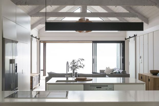 Bright open-plan kitchen and living area with a large white island, integrated appliances, timber cabinetry, expansive sliding glazing, and a vaulted ceiling with exposed white-painted beams and clerestory windows. A linear black pendant light hangs above the island, while the living room is visible beyond.