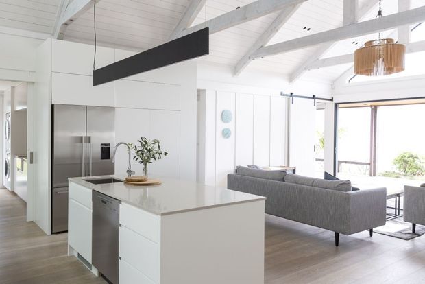 Bright open-plan kitchen and living area with a white island, integrated stainless-steel appliances, grey sectional sofa, light timber flooring, white-painted exposed ceiling beams, and large glazed doors opening to the outdoors. A linear black pendant and woven timber pendant add contrast.