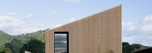 Exterior view of a minimalist contemporary building set against a wooded, hilly landscape. The facade is largely covered in closely spaced vertical timber slats, creating a strong screen-like texture and a sharply rising roofline or parapet. A tall, dark-framed glazed opening punctuates the otherwise solid facade.