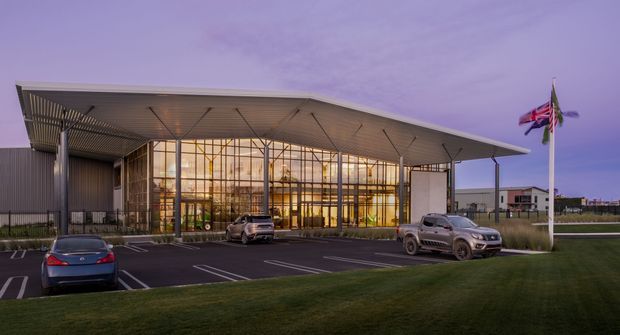 A contemporary commercial building viewed from across a landscaped lawn and parking area at dusk. The main facade features a broad, shallow projecting roof canopy, extensive floor-to-ceiling glazed curtain walling, and dark metal cladding. Interior lighting glows through the transparent entrance or lobby volume. Flagpoles and parked vehicles are visible outside, but the building is the dominant subject.