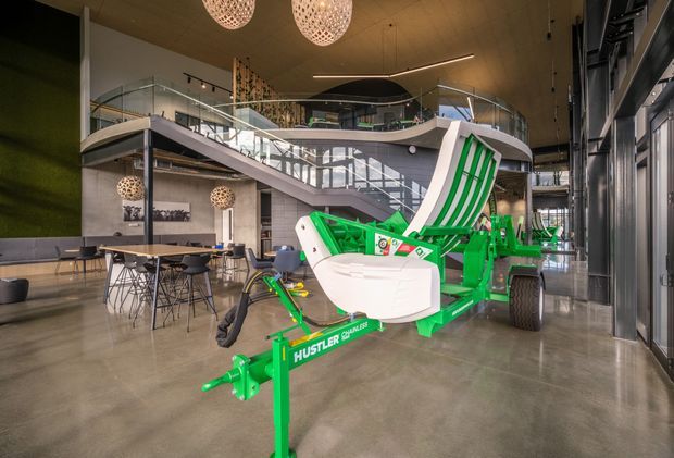 A spacious contemporary commercial showroom or office interior with polished concrete flooring, a double-height atrium, mezzanine walkway, glass balustrades, exposed structural elements, and large industrial-style glazing. A bright green agricultural implement is prominently displayed in the foreground. The space includes a small café-style seating area, sculptural pendant lights, and a large green feature wall.