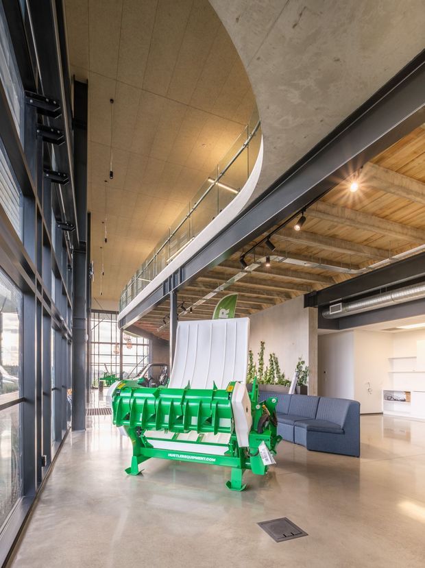 A bright, double-height commercial or showroom interior with polished concrete flooring, expansive black-framed glazing, exposed timber roof structure, exposed steel beams and ducts, and a curved mezzanine with glass balustrading. A large green agricultural machine is displayed in the foreground, with a lounge seating area and minimal white kitchenette or joinery visible to the side.