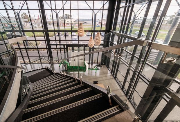 A dramatic multi-level interior stairway viewed from above, featuring broad dark carpeted stairs with metallic edge trims, stainless-steel handrails, extensive black-framed floor-to-ceiling glazing, exposed structural framing, and several woven pendant lights. A lower-level lobby with green seating or display elements is visible below, while streetscape and landscaping can be seen through the glass.
