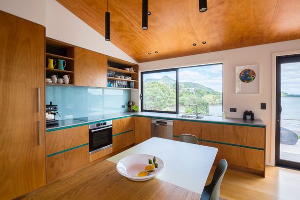 Contemporary open-plan kitchen and dining area with warm timber cabinetry, a matching timber-lined ceiling, turquoise glass splashback, integrated appliances, and a white dining table with dark chairs. Large windows and a glazed door provide views toward a leafy hillside and waterfront beyond. Black linear and recessed lighting add contrast to the warm natural finishes.