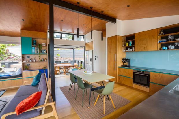 Open-plan kitchen and dining area with extensive warm timber cabinetry and ceiling lining, a bright turquoise glass splashback, integrated oven and cooktop, and a central dining table with green chairs. The room opens to a living area with built-in shelving and a sofa, while large glazed doors connect to an outdoor courtyard. Black steel structural posts and beams, pendant lights, and polished timber flooring create a contemporary, warm interior.